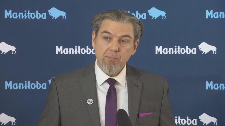 Man in grey suit, purple tie and white shirt stands in front of a mic. His hair is grey. 