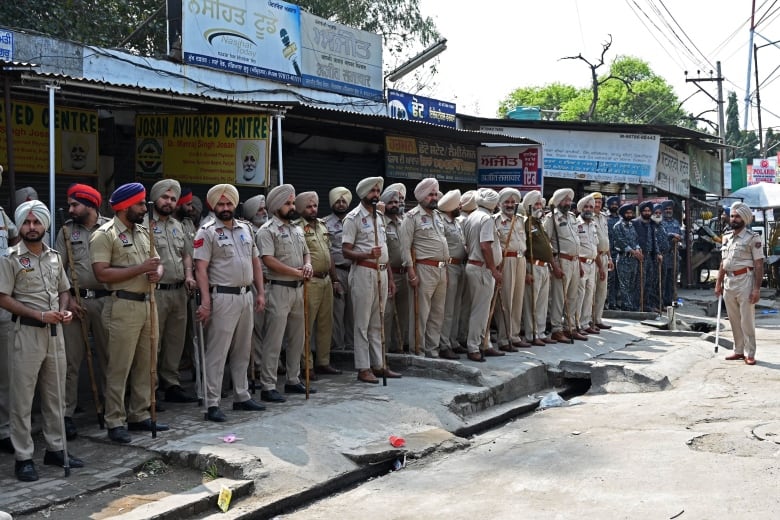 A line of police officers, wearing khaki uniforms and matching turbans, stand in front a row of shops. 