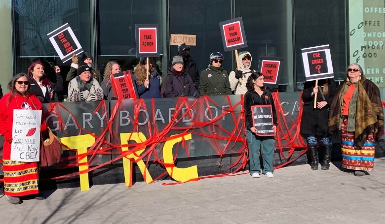 A group of about 20 people stand before a sign marking the Calgary Board of Education Centre, which protesters lined with red tape. 