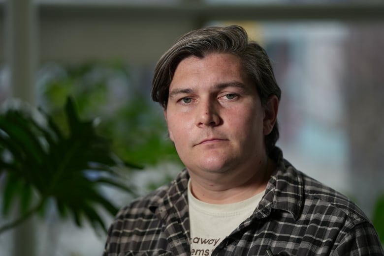 A man wearing a plaid shirt over a T-shirt with a serious expression looks at the camera