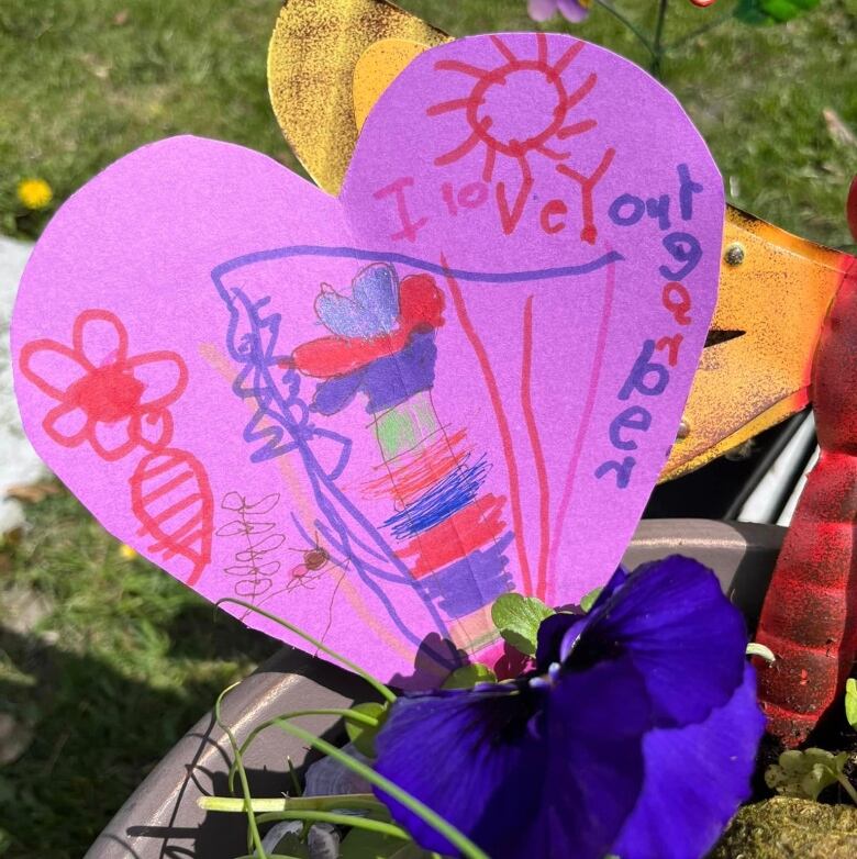 Photo of a hand made note shaped as a heart on purple craft paper with the message, 'I love you grandpa' written in children's handwriting along with hand drawn designs is placed in a floral planter.