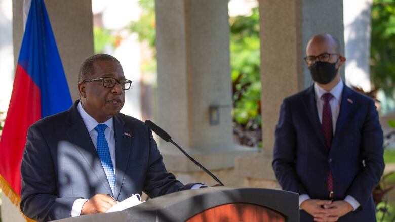 A man speaks at a podium while another man, wearing a mask, listens in the background.