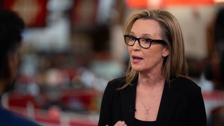 A middle-aged white woman with long, straight, sandy blonde hair, gestures with her hand as she talks to an interviewer, who can be seen in profile to the left of the frame.