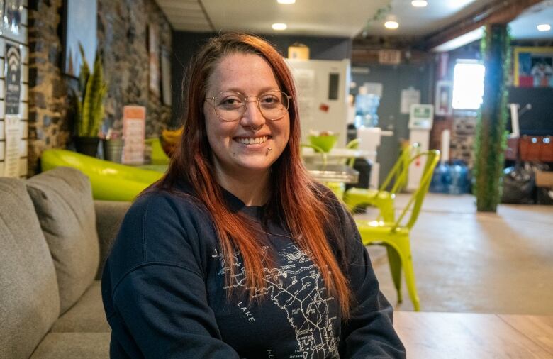 A young woman with red hair and glasses sits on a couch, smiling.