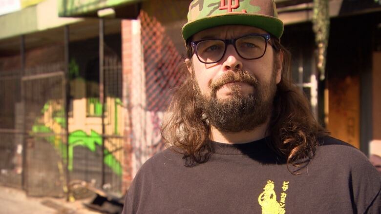 A man with a beard and long brown hair wearing purple-framed glasses and a camouflaged baseball hat stands for a news interview.