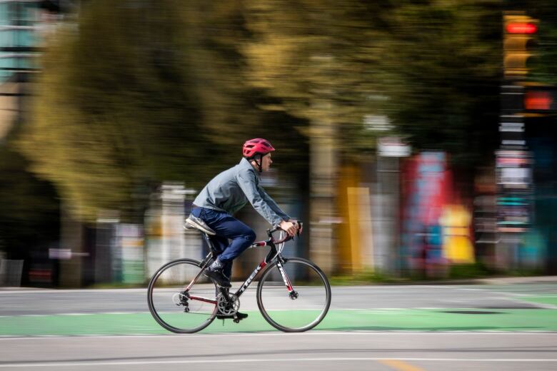 A man wearing blue pants and a blue fleece pull-over rides a red and black bike through downtown Vancouver. The cyclist is in clear focus while the city landscape is blurred in the background.