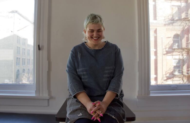 Non-binary person with short blond hair sits on stool in studio. 