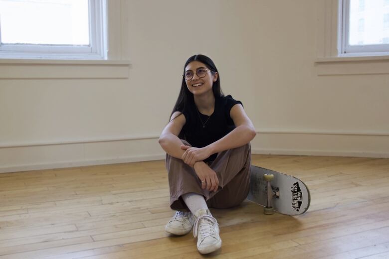 Non-binary person with long, brown hair sits on hardwood floor beside their skateboard.