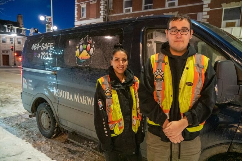 Two people wearing fluorescent vests stand in front of a black van.