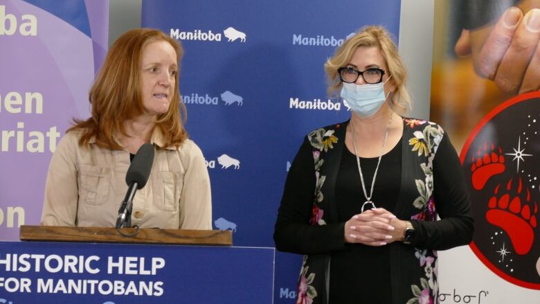 A woman standing at a podium, next to another woman wearing a surgical mask.