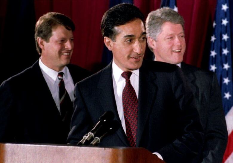 A man in a suit and tie is shown at a podium, with two other similarly dressed men seen in the background.