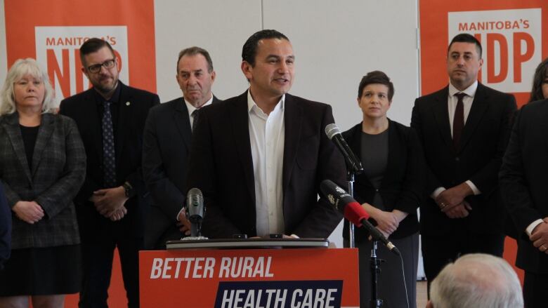 A man in a white collared shirt and dark blazer speaks to a crowd at an election campaign announcement.