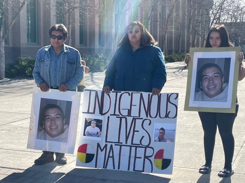 Three members of an Indigenous family stand outside a building holding two signs, each with a photograph of a smiling young man, while a third sign with more pictures of the same person on it reads: Indigenous lives matter.