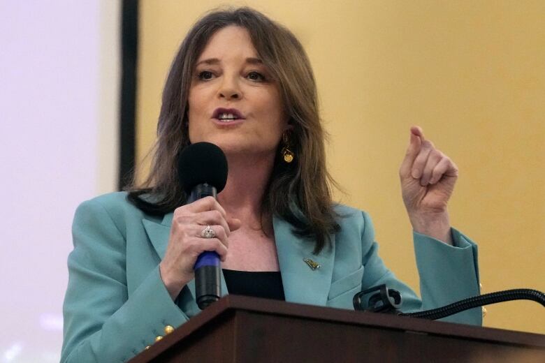 Woman with shoulder length brown hair wearing a light blue jacket, gestures as she speaks into a microphone