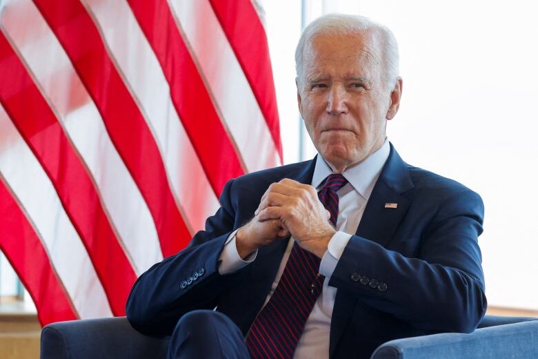 White-haired man in a blue suit, seated in front of a U.S. flag