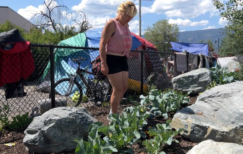 A woman in a pink vest and a black short stands near vegetables on the ground, with fencing behind her.