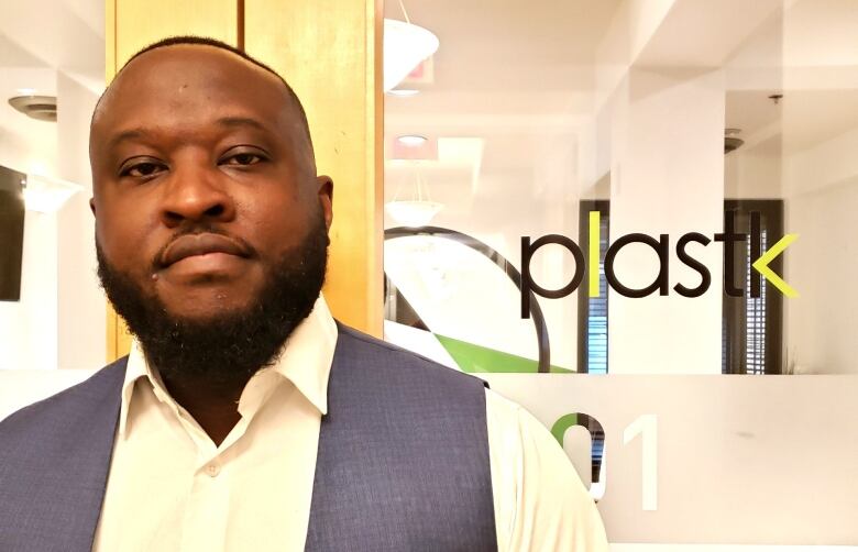 A bearded man wearing a white collared shirt and blue vest faces the camera with a slight smile. He is standing in from of a pair of glass doors printed with the Plastk logo in green and black text. 