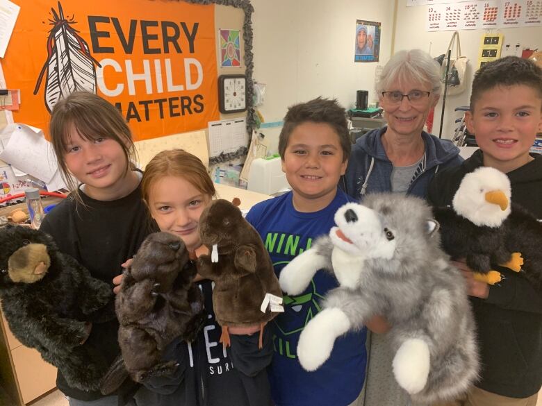 Four children hold up stuffed animals in a classroom with woman in glasses and white hair. In the background is an orange flag that says, 