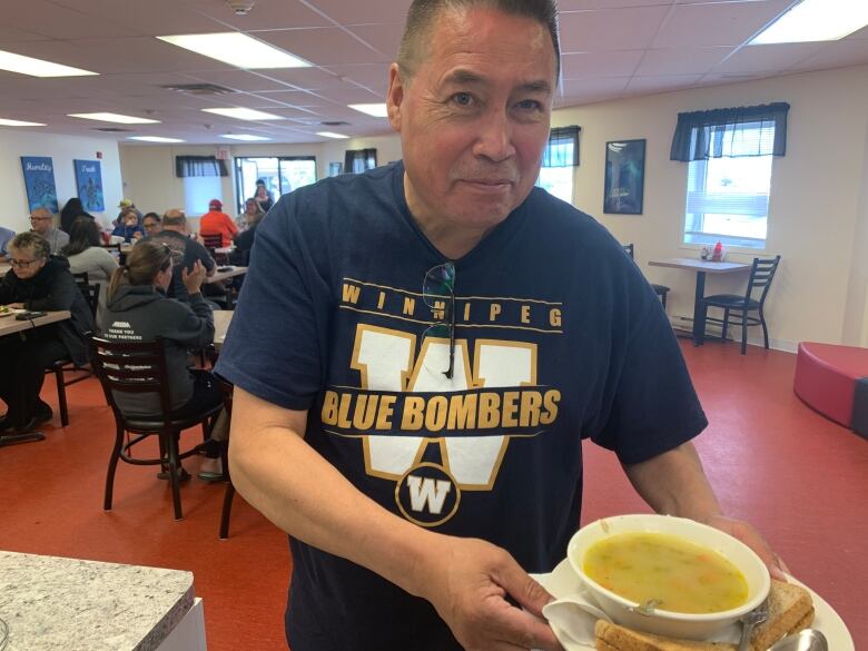 A man in a Winnipeg Blue Bombers t-shirt holds a plate with a bowl of soup and a sandwich on it in a restaurant.