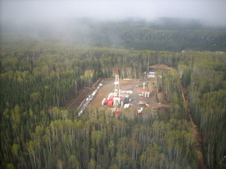 In the middle of a thick, misty forest, an aerial shot shows us an area of clear cut with oil and gas infrastructure and a tall well head. 