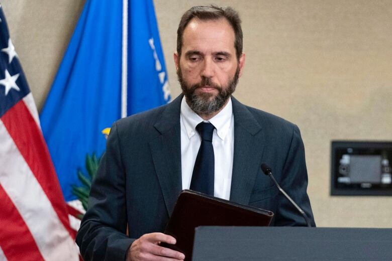 A man in a suit and tie is shown leaving a podium, holding a leather folder.