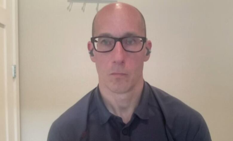 A bald man in glasses and a black button down shirt looks at the camera with a straight face.