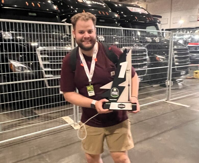 A man poses for a photo holding a trophy, wearing a burgundy shirt and khaki shorts