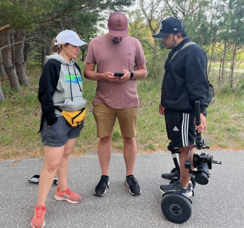 A woman standing in a park with two men. One man has a camera and is balancing on a Segway.