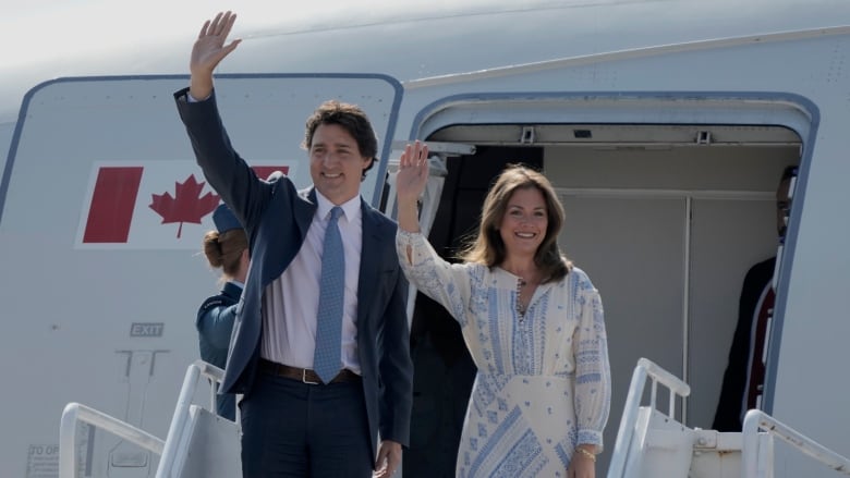 A man and a woman stand on a stairway outside the entrance to an airplane and wave.