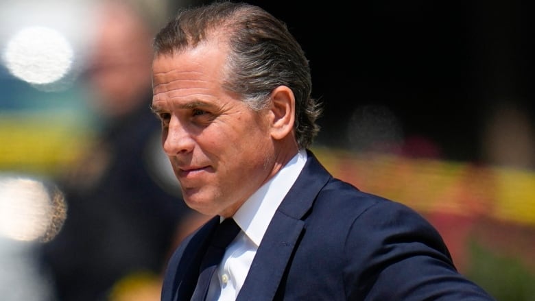 A man with brownish-greyish hair wears a navy suit and tie.