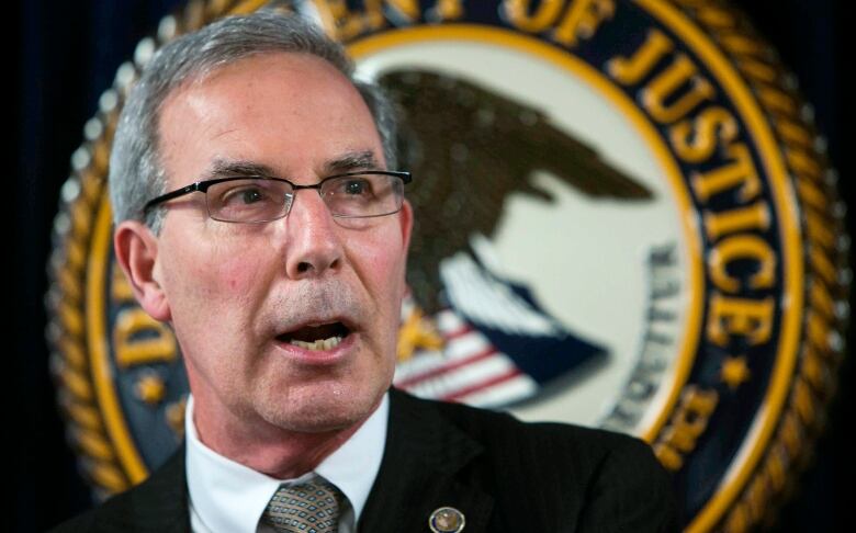 A man with grey hair, wearing glasses and a black suit, speaks while standing in front of the seal for the Department of Justice.