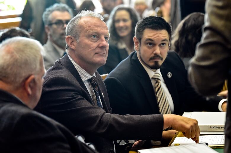 Two men wearing suits and ties look up intently, as one points to a binder on the table.