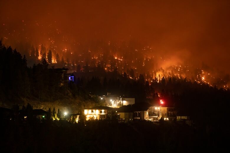 Well-lit mountainside homes are pictured in the foreground, and behind them, a dark night sky lit orange by burning wildfires.