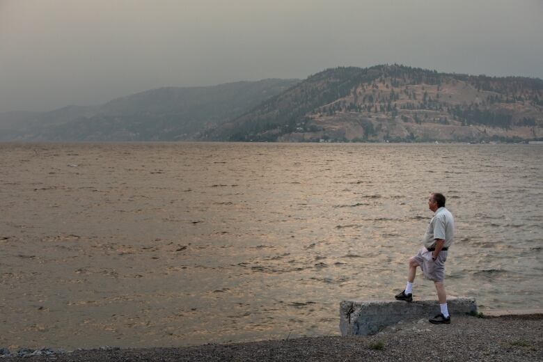 A man rests his leg on a stone overlooking a smoky lake.