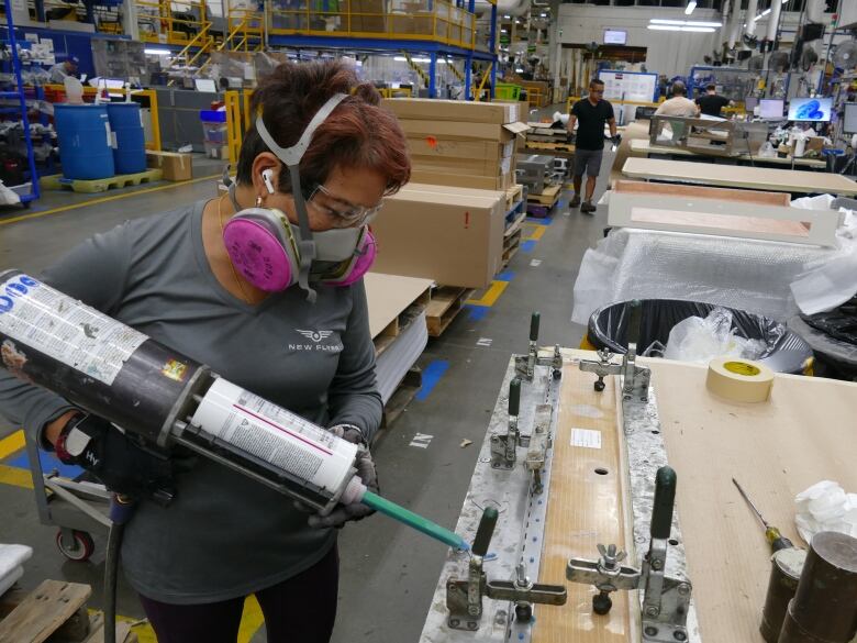 A woman wearing a ventilator mask stands in a factory squeezing gel from a large applicator onto a machinery part.