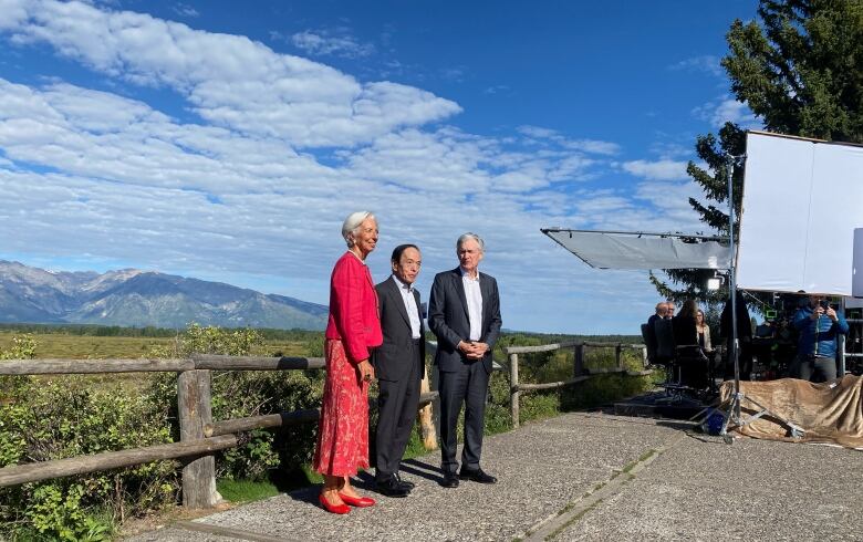 A woman with short white hair, wearing a red outfit, stands beside two men in grey suits with mountains in the background.