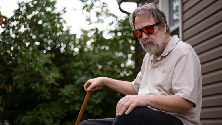 An old man with a beard sits on stairs outside a small house.