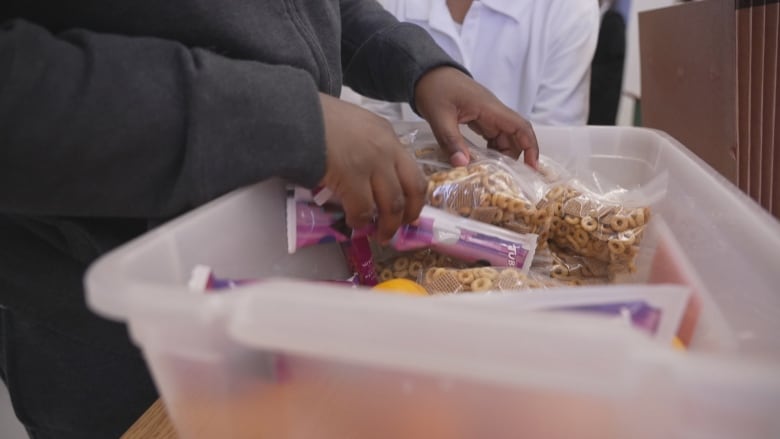 A young person's hands are shown rifling through a snack bin filled with ziplocks of cheerios, oranges and tubes of yogurt.