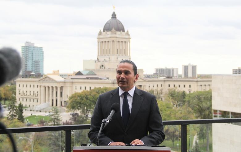 A man in a suit and tie stands on a balcony, overlooking the Manitoba Legislative Building in the background.