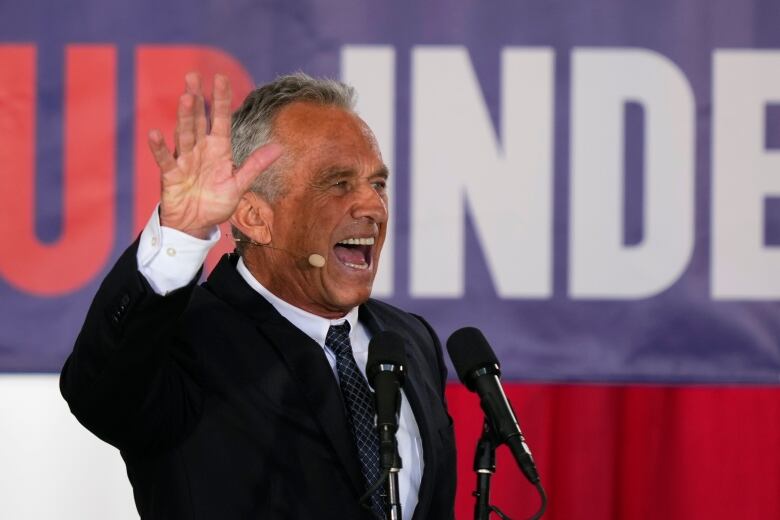 A man in a suit and tie raises his hand while speaking on a stage.