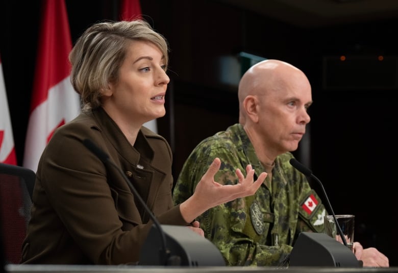 A woman seated at a table gestures while speaking. A man in uniform is seated next to her. 