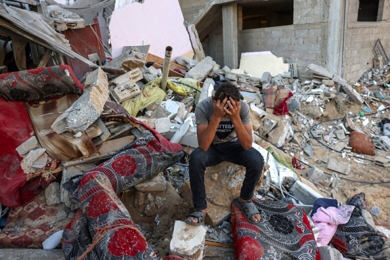 A young man sits on a stone with his head in his hands amid a pile of rubble.