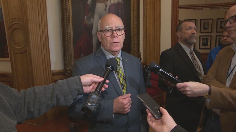 A bald man with glasses speakers into several microphones held out by reporters.