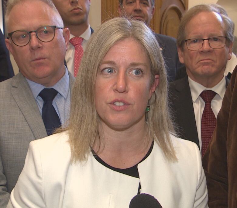 A woman in a white blazer speaks into a microphone while surrounded by people.