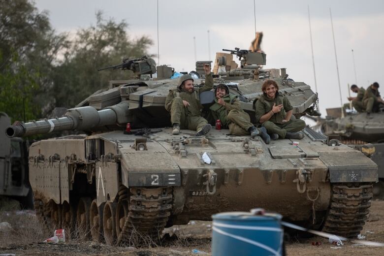 Three young men in military gear sit casually and smile on a large, green tank.