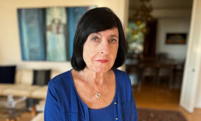 An aged white woman with a black bob and blue cardigan sits in her living room 