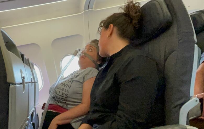 Looking down an airplance row, a woman with a ventilator looks uncomfortable sitting next to her support worker.