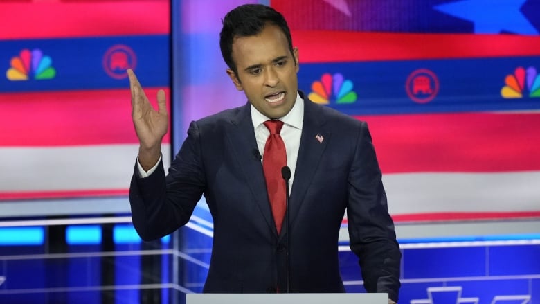 A man, wearing a dark blue suit, white shirt and red tie, speaks at a microphone with his right hand raised in a gesture. 