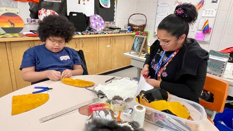 A woman and a child are sitting at a table working on moccasins