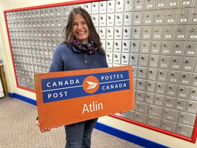 A woman stands by a wall of post offfice boxes, holding a Canada Post sign reading 'Atlin.'